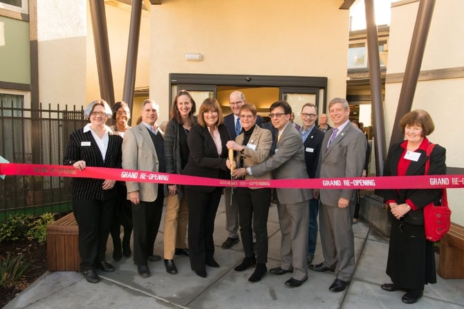 Group cutting a ribbon with big scissors. The ribbon reads "Grand Re-Opening"