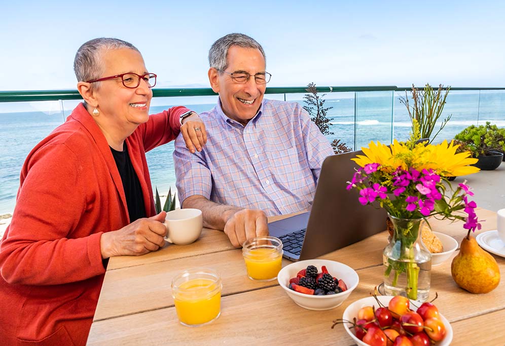 Janet and Mark enjoying breakfast and looking at a laptop on a balcony with a scenic ocean view