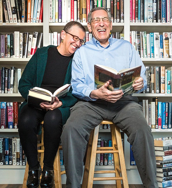 Janet and Mark sitting on stools and smiling while holding open books